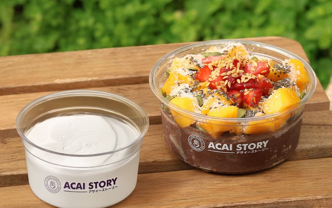 An Acai Story acai bowl topped with mango, strawberries, and seeds beside a yogurt cup