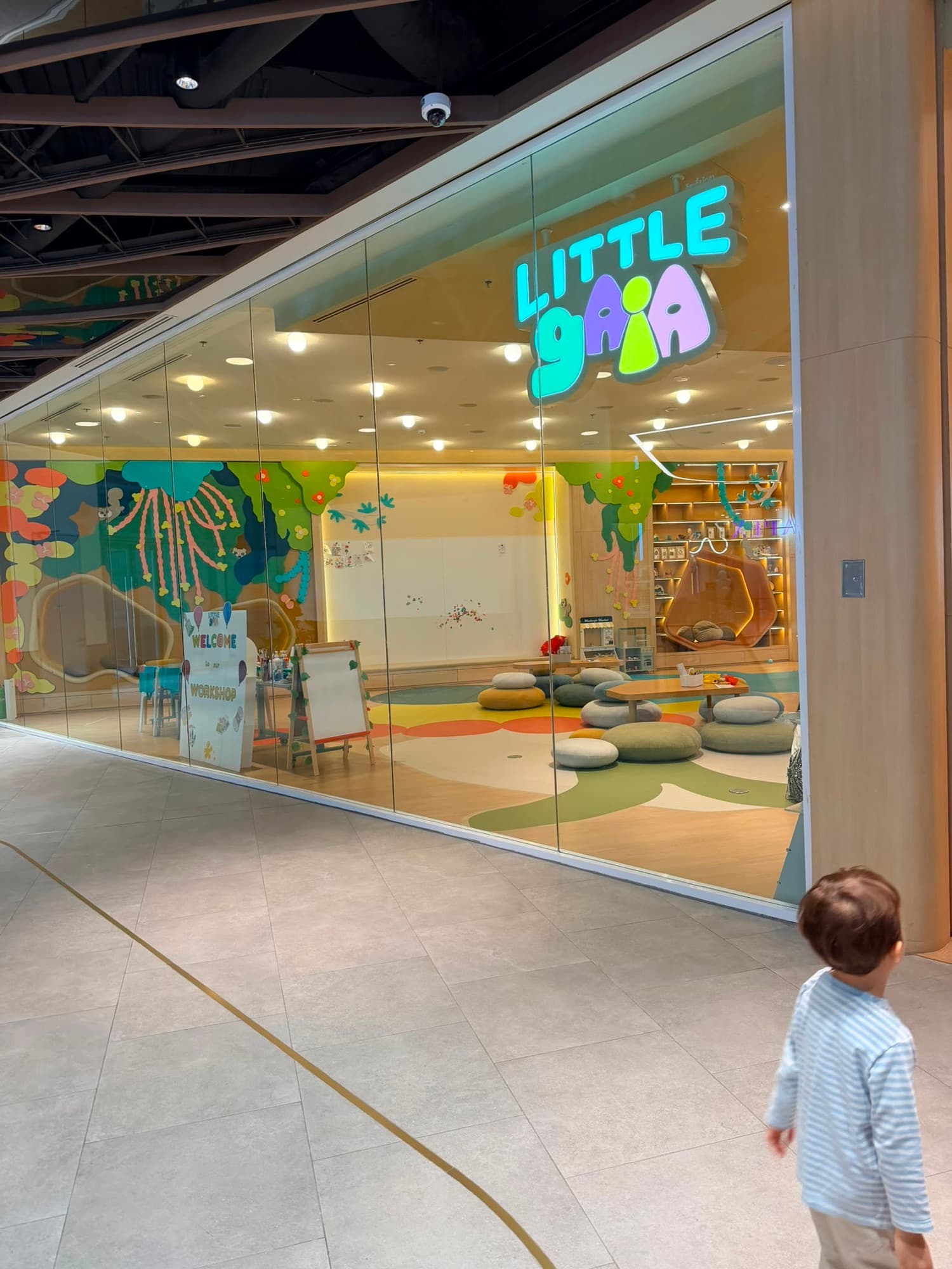A young boy walking toward the glass entrance of Little Gaia, a pastel, nature-themed kids play space inside One Bangkok mall in Bangkok