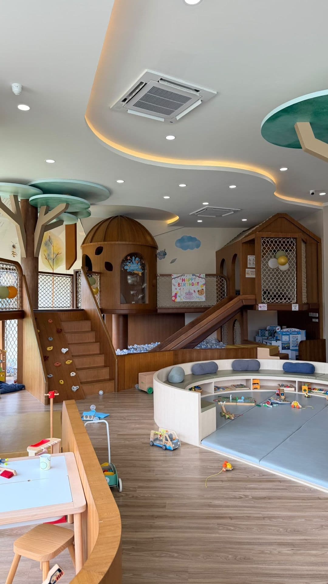 A wooden, nature-inspired indoor playground at Little Owly's creative nest and café in Bangkok, with treehouse structures, a small climbing wall, and a wooden slide into a ball pit