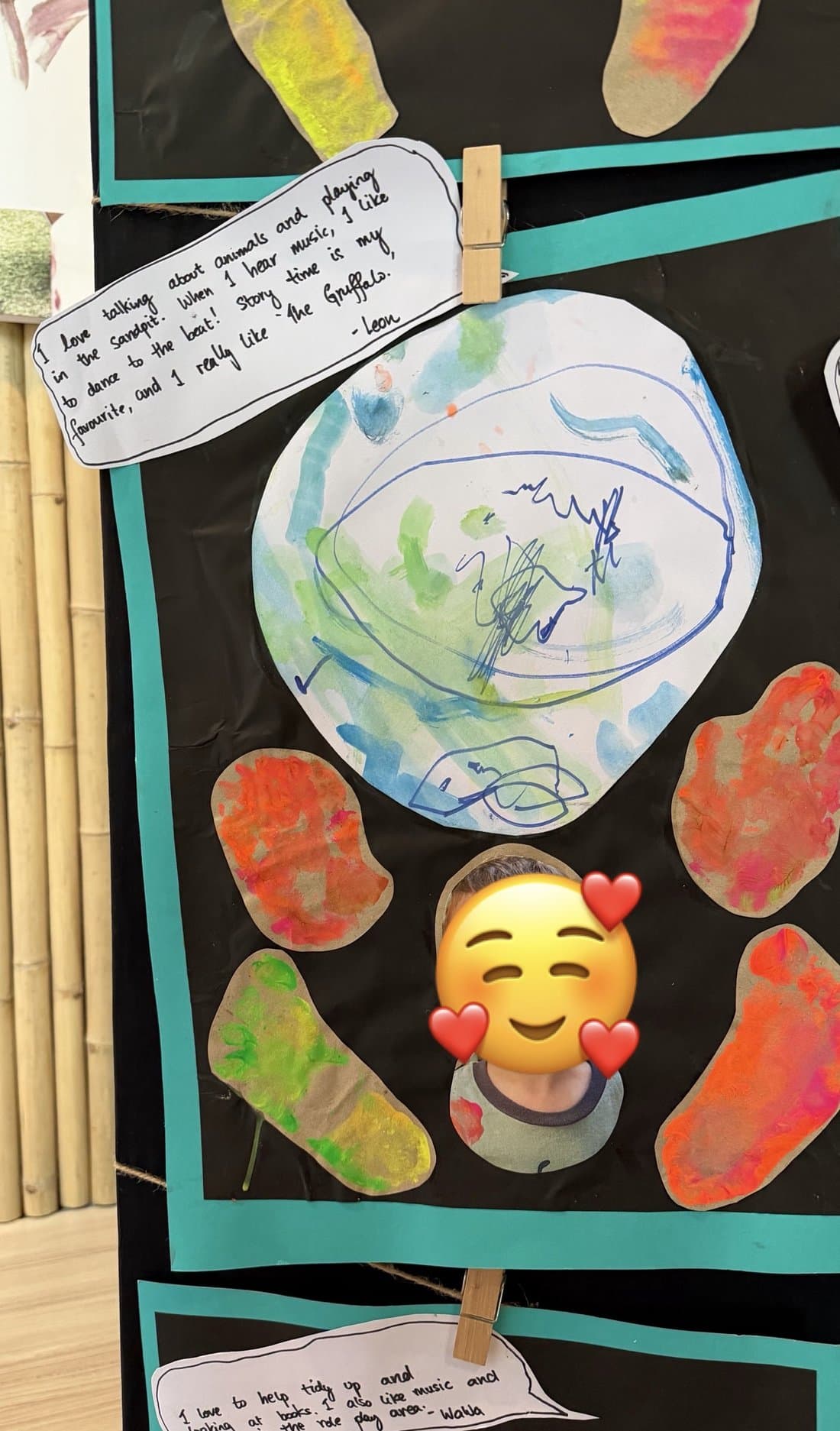 A classroom display board with a child’s round watercolor painting in blues and greens at the center, surrounded by abstract orange and red cutout artworks, with handwritten notes pinned at the top and bottom