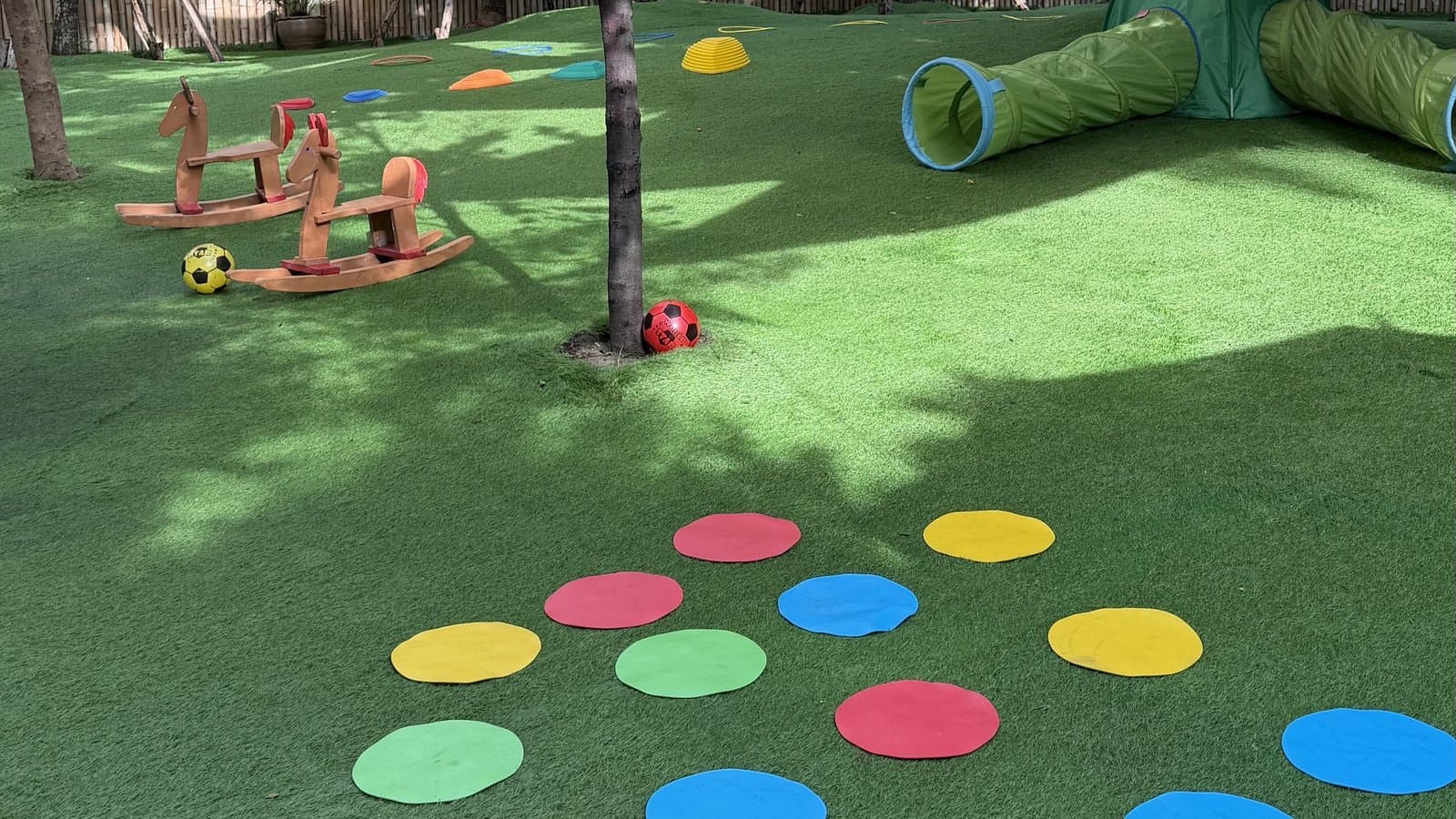 A Reggio-inspired preschool outdoor playground in Bangkok with shade sails overhead, rainbow stepping circles on artificial grass, wooden rocking horses, and a play tunnel