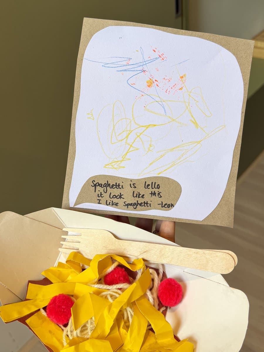 A child’s spaghetti craft made from yarn, red pompoms, and yellow paper strips in a small cardboard takeaway box