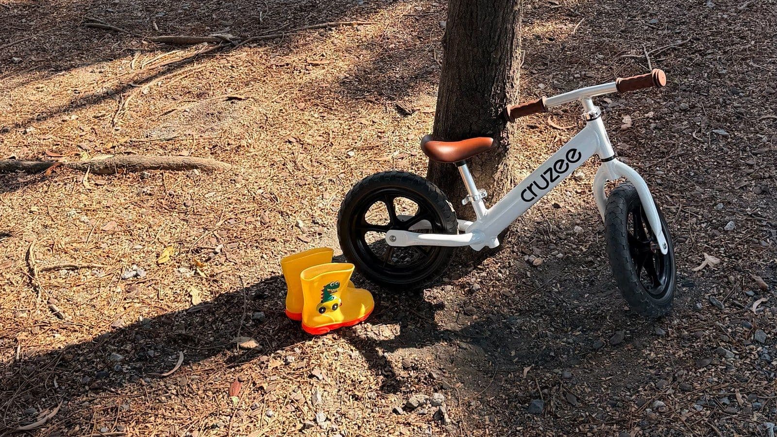 A small balance bike resting against a tree at Nong Bon Lake Park in Bangkok, with yellow rain boots on the ground and the lake behind through the trees