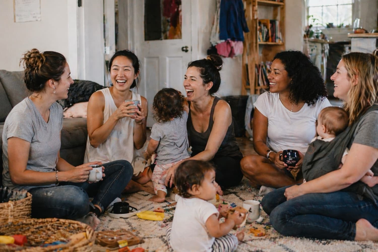 A circle of moms laughing together in a living room while their toddlers play around them