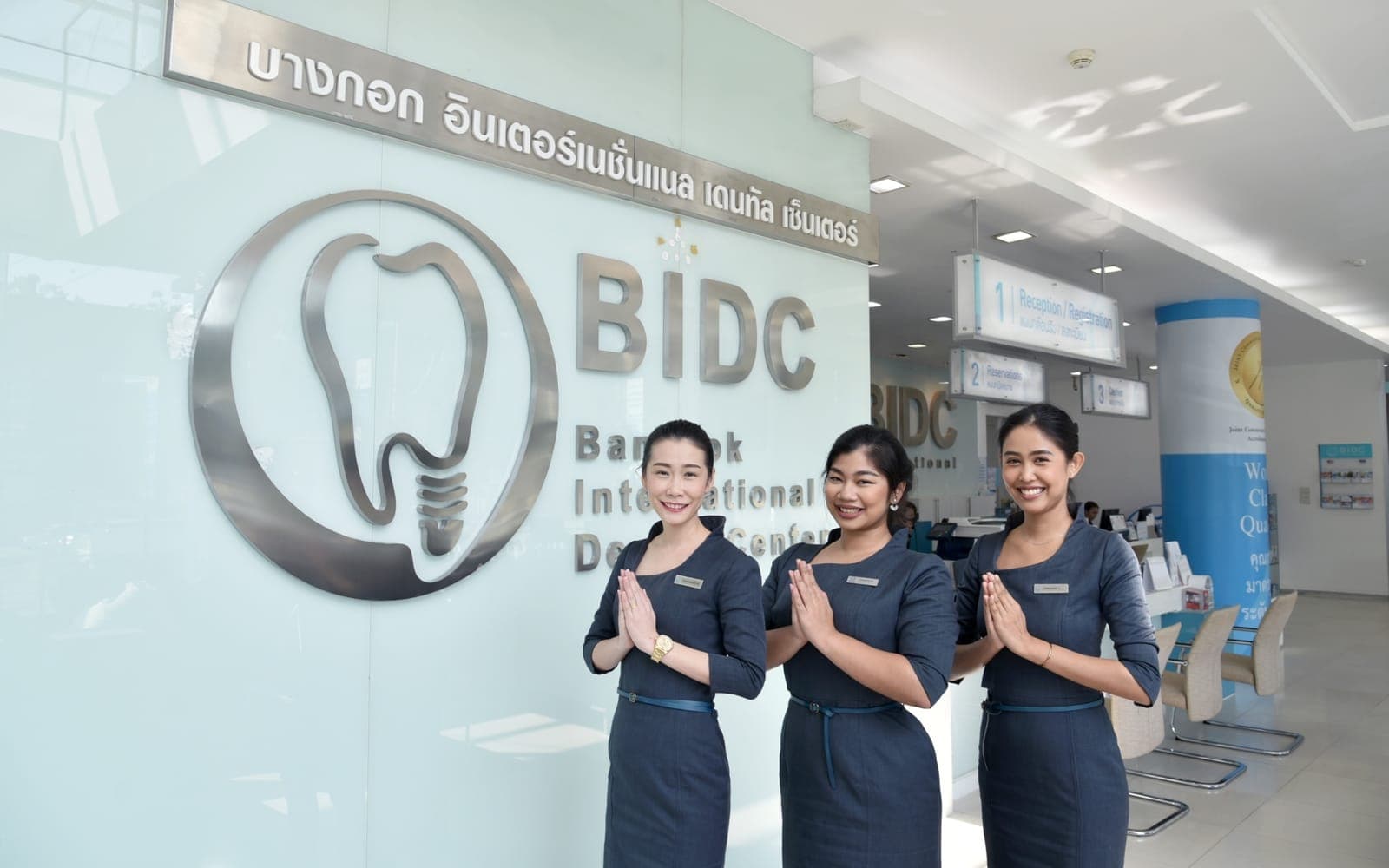 Three staff greeting visitors with a wai beside the BIDC logo at a Bangkok International Dental Center reception, Bangkok