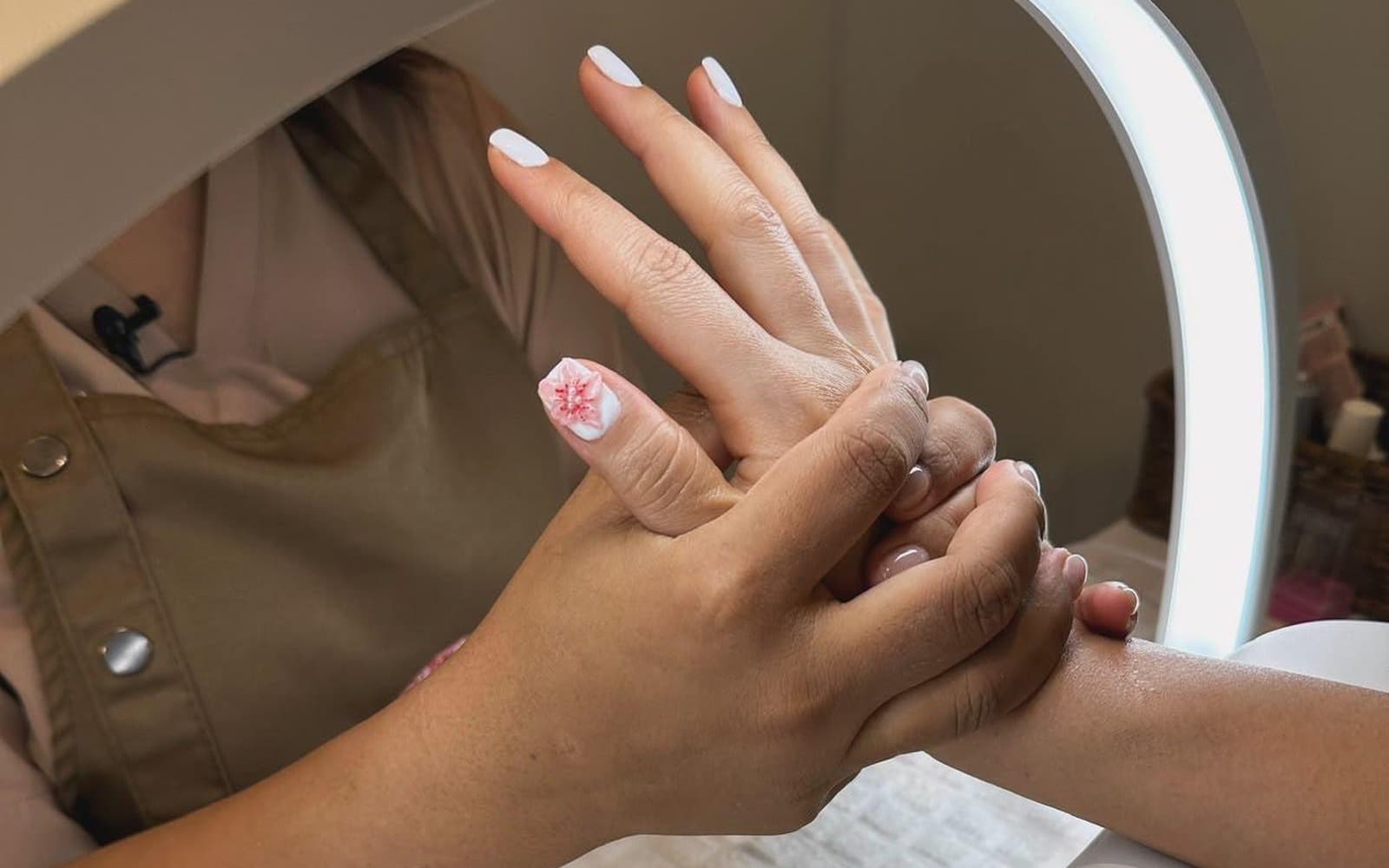 A manicure in progress at Olala Glowhaus, a technician massaging a hand under a ring light, with white nails and a pink flower design