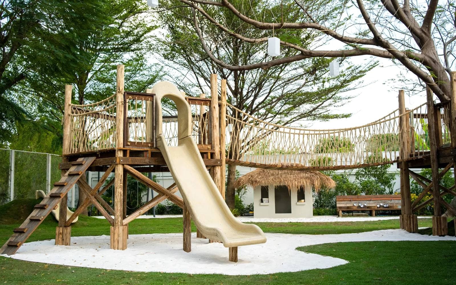 The wooden playground at Sweet Poppy, with a slide, rope bridges, and a white sand pit under tall garden trees