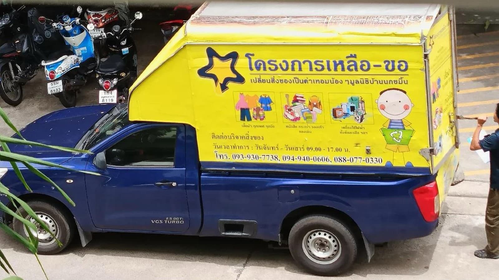 The bright yellow Leua-Kho donation truck, a blue pickup with a yellow cargo box covered in the project star logo and Thai text, parked at the curb while a volunteer opens the back
