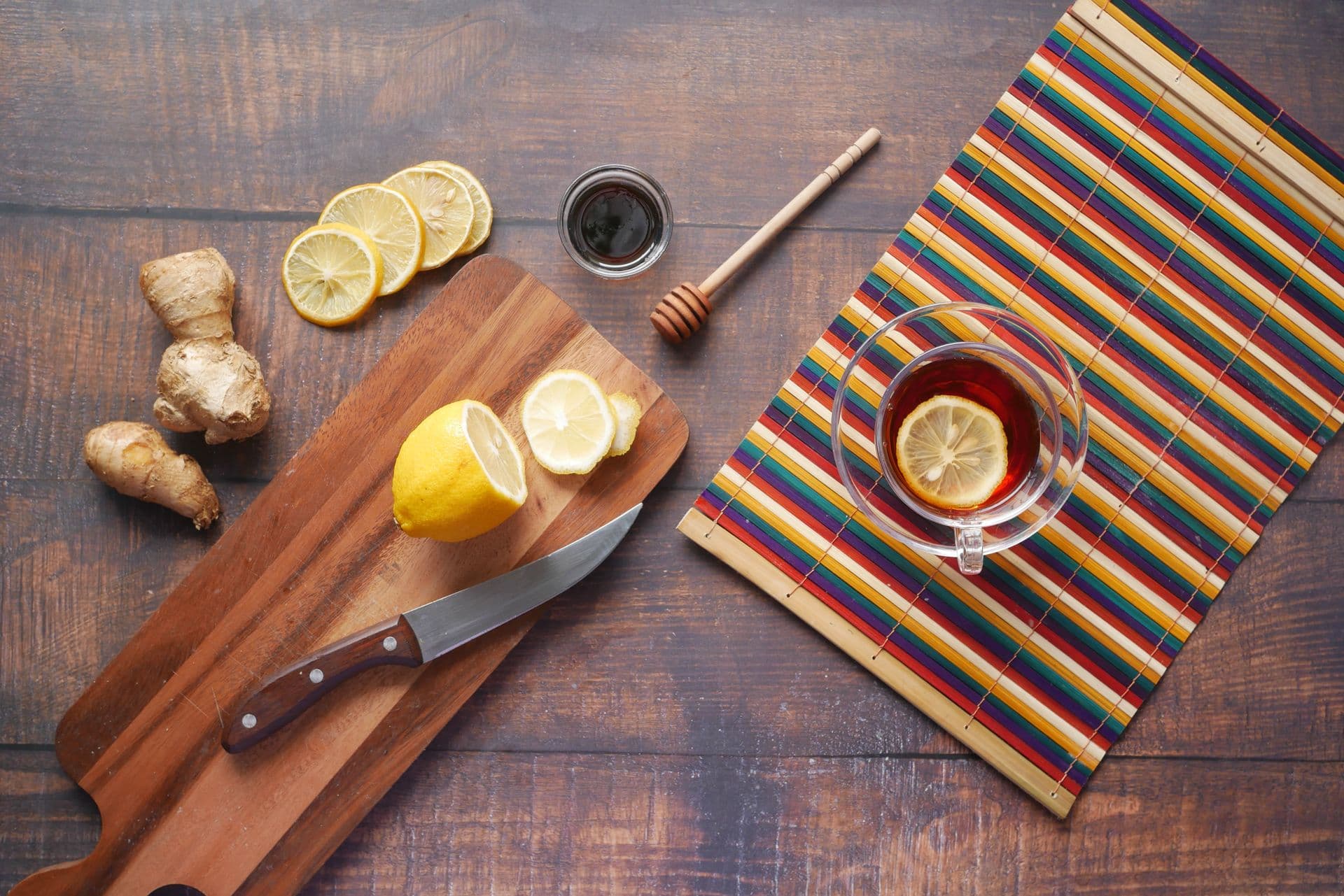 A flat lay of natural home remedies on a wooden board: fresh ginger root, sliced lemon, a small jar of honey with a wooden dipper, and a cup of warm ginger tea